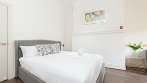 A neatly made bed with a grey headboard, white bedding, and a single white towel on the footboard. A framed abstract painting hangs above the bed, and a vase with yellow flowers is on a side table to the right. - Stunning Sydney Home 9 (Millers Point)