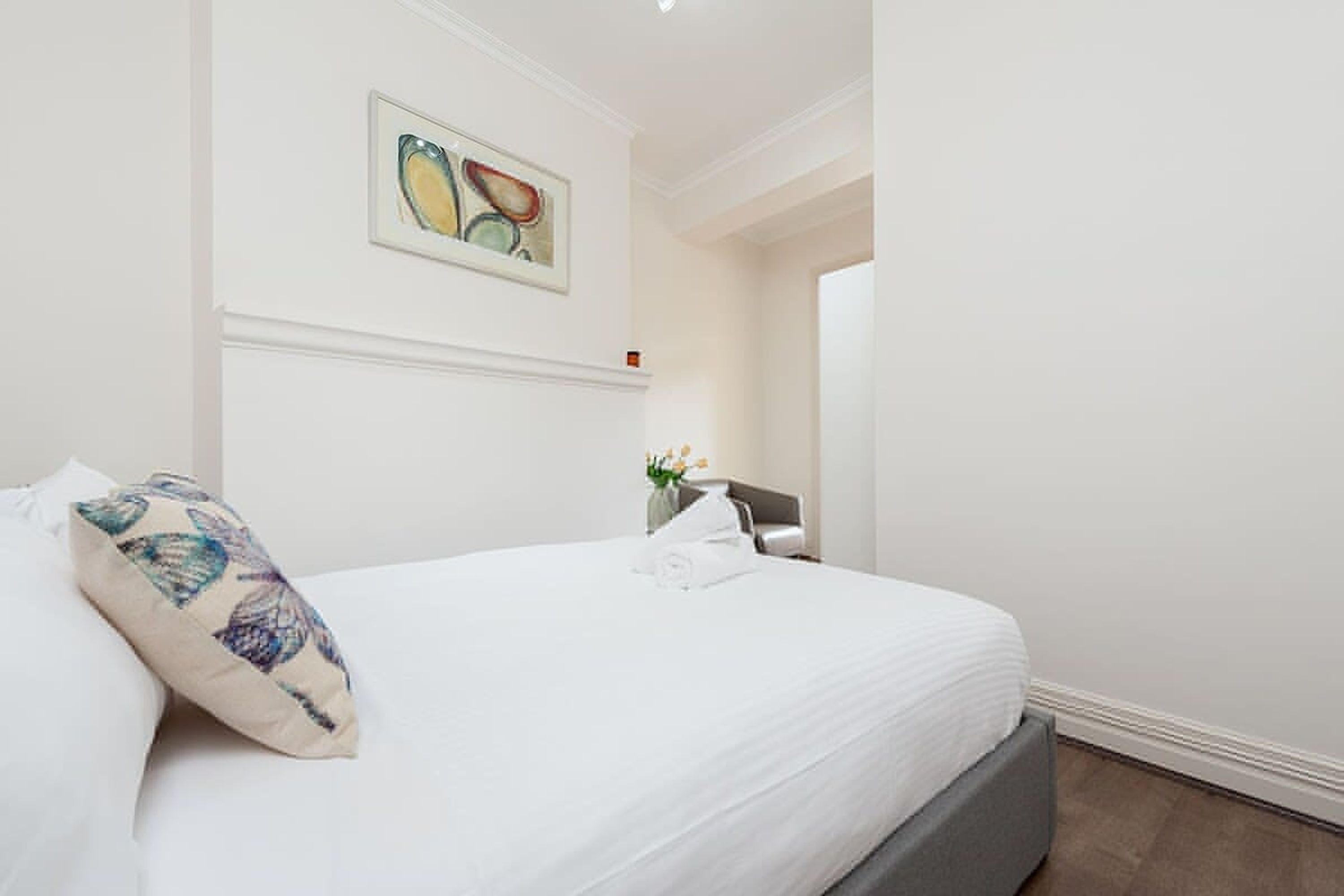 A neatly made bed with a floral pillow, a small vase with flowers, and a painting on the wall.