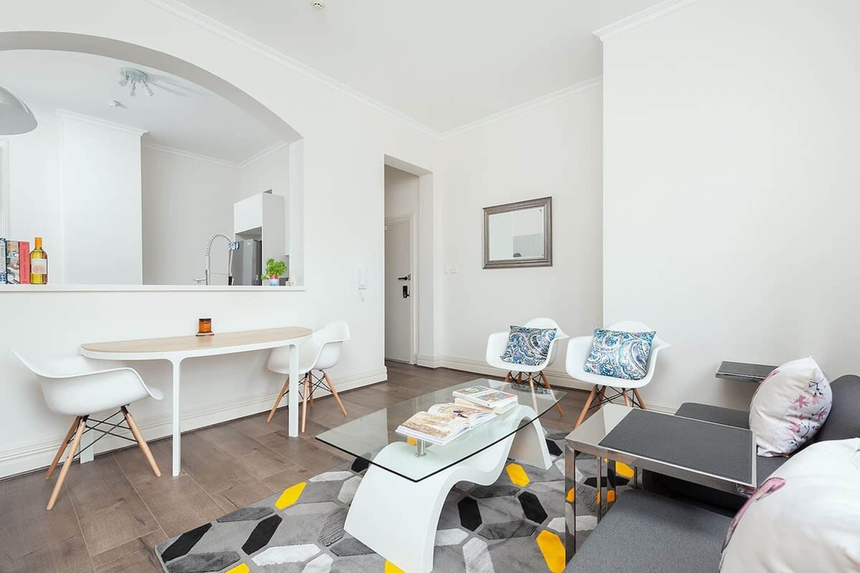 A modern living room with a glass coffee table, white sofas, and a dining area with a white table and chairs.