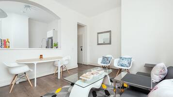 A modern, well-lit room with a glass-top table, two white chairs, a sofa, and a small coffee table.