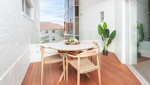 A balcony with a round wooden table and chairs, a potted plant, and a view of buildings in the background. - Ultimate Bondi Living (Bondi Beach)