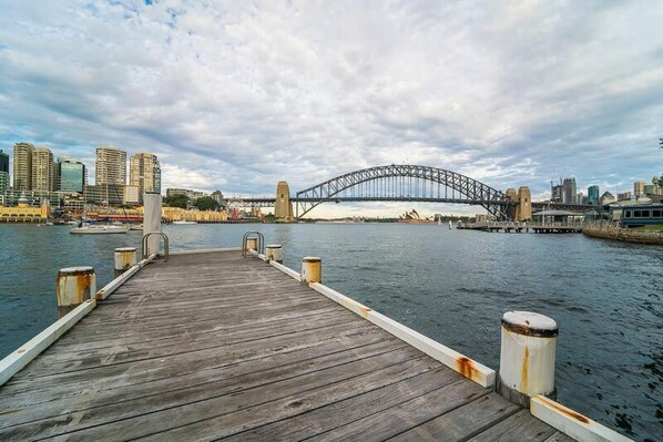 A wooden pier leading to a bridge over water with city buildings in the background. - Harbourside 76 in North Sydney (McMahons Point)