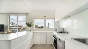 A modern kitchen with white cabinetry, a built-in oven, and a dishwasher. There are two windows offering views of a city skyline and a bridge. - Harbourside 49 in North Sydney (McMahons Point)