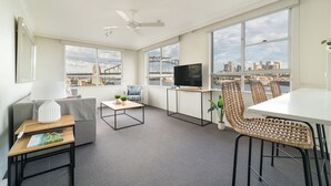 A modern living room with a TV, seating, and large windows offering city views. - Harbourside 49 in North Sydney (McMahons Point)