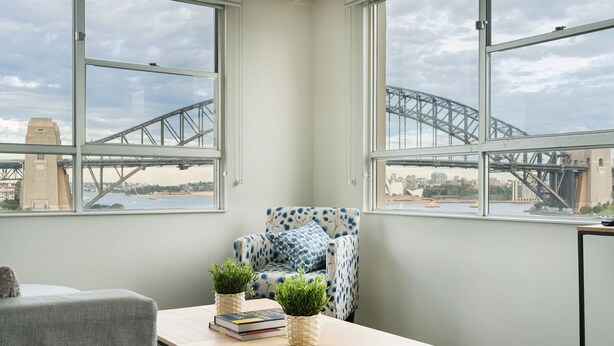 A room with a view of a bridge, a patterned armchair, a small table with books and plants, and a grey sofa. - Harbourside 49 in North Sydney (McMahons Point)