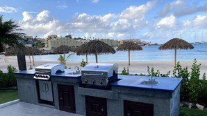 An outdoor kitchen with a grill and a built-in sink, overlooking a beach with umbrellas and a view of the ocean. - Beachfront, ground level condo, steps away from beach, pool and relaxation! (Cole Bay)