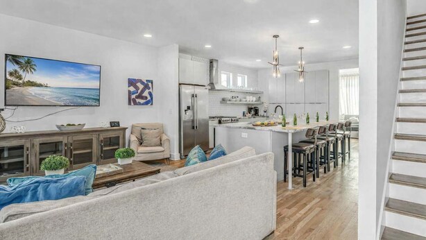 A modern living room with a sofa, a coffee table, and a TV. There is a kitchen area with a refrigerator, a dining table with chairs, and a kitchen island.