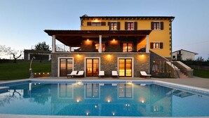 Una casa de dos pisos con piscina, paredes de piedra y un balcón con luces.