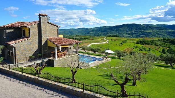 Un edificio de piedra con tejado de tejas, una piscina y vistas a colinas verdes y una carretera sinuosa.
