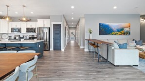 A modern kitchen with stainless steel appliances, a dining area with a wooden table and chairs, and a living room with a sofa and a large landscape painting. - Large-group Retreat w/Hot Tub, 2 Kitchens, Grill (Branson)