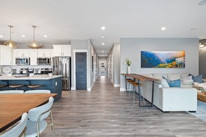 A modern kitchen with stainless steel appliances, a dining area with a wooden table and chairs, and a living room with a sofa and a large landscape painting. - Large-group Retreat w/Hot Tub, 2 Kitchens, Grill (Branson)