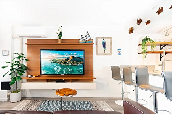 A modern living room with a large flat-screen TV, a wooden entertainment unit, a dining area with a bar stool, and a potted plant.
