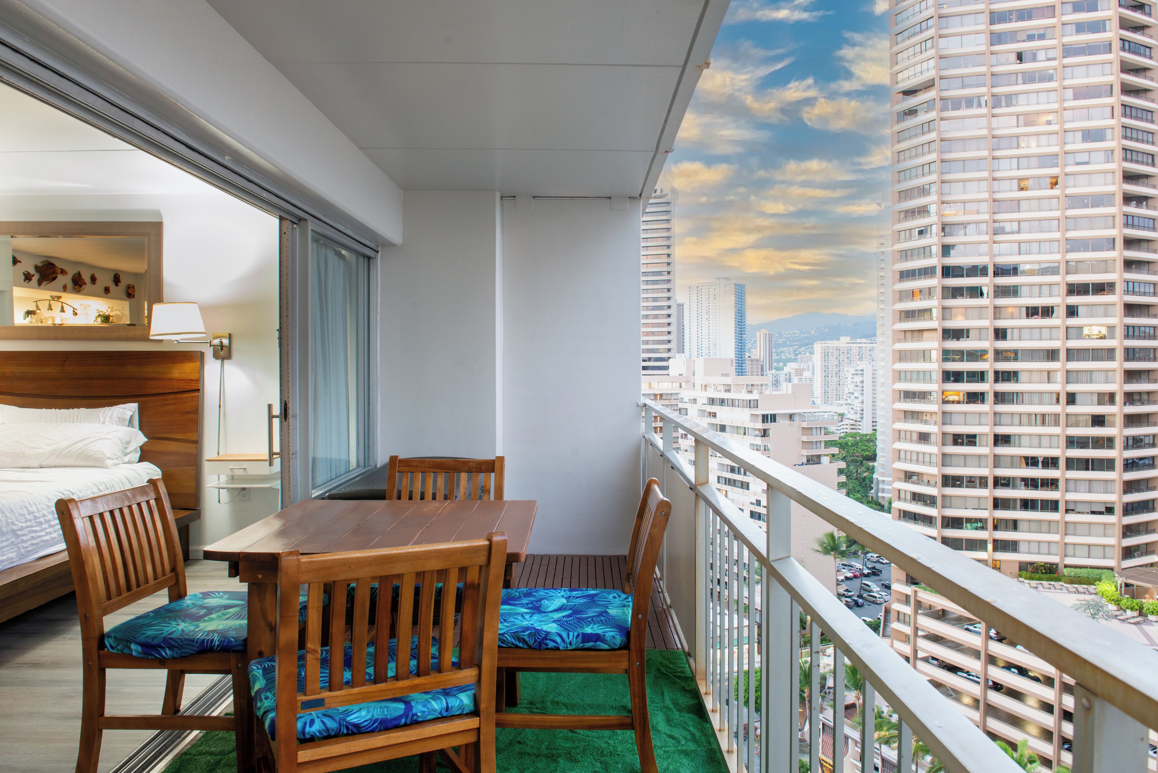 A balcony with a wooden table and chairs, a bed, and a view of a cityscape.