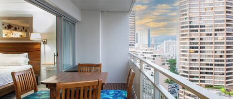 A balcony with a wooden table and chairs, a bed, and a view of a cityscape.