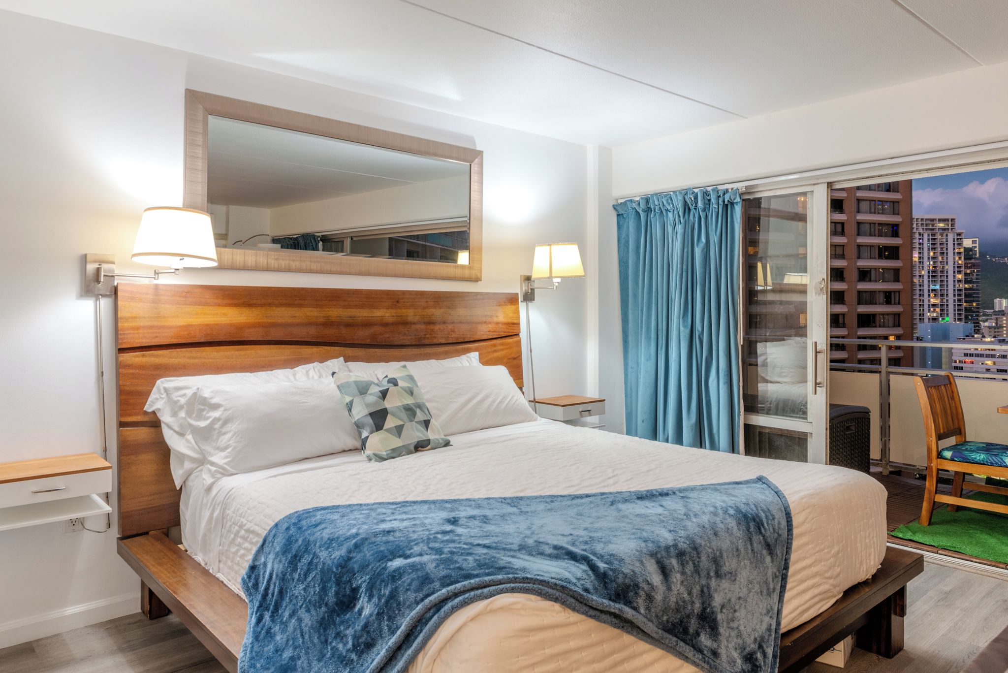 Een hotelkamer met een bed, een spiegel boven het bed, een nachtkastje, een lamp, een raam met gordijnen en uitzicht op de stad.