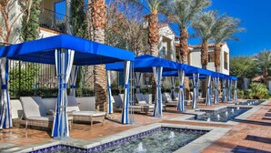 A poolside area with lounge chairs and umbrellas, surrounded by palm trees and a building with a balcony. - Stunning 2bed/2bath Villa - Amazing Location+Views (La Quinta)
