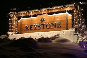 Et træskilt med ordet "KEYSTONE" oplyst af lys, omgivet af sne og festlige pynt.