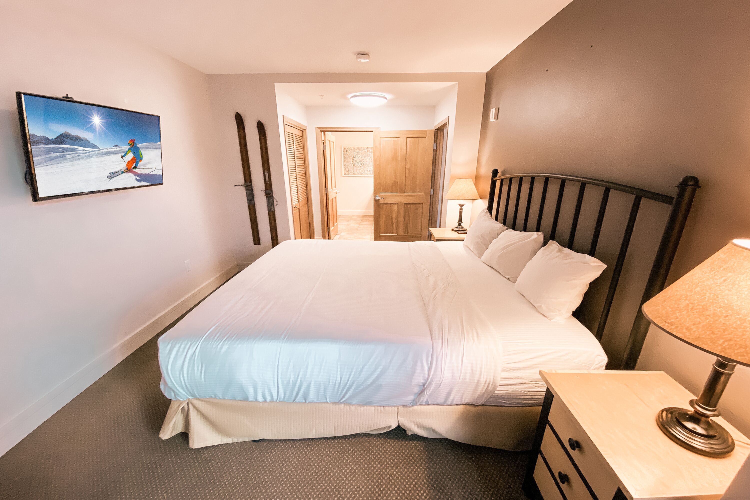 Une chambre d’hôtel avec un grand lit, une table de chevet avec une lampe, un téléviseur fixé au mur et une porte donnant sur une autre pièce.