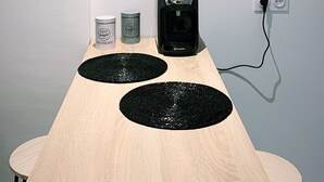 Et minimalistisk rum med et træbord, to sorte underkopper, en kaffemaskine og en lille potteplante.