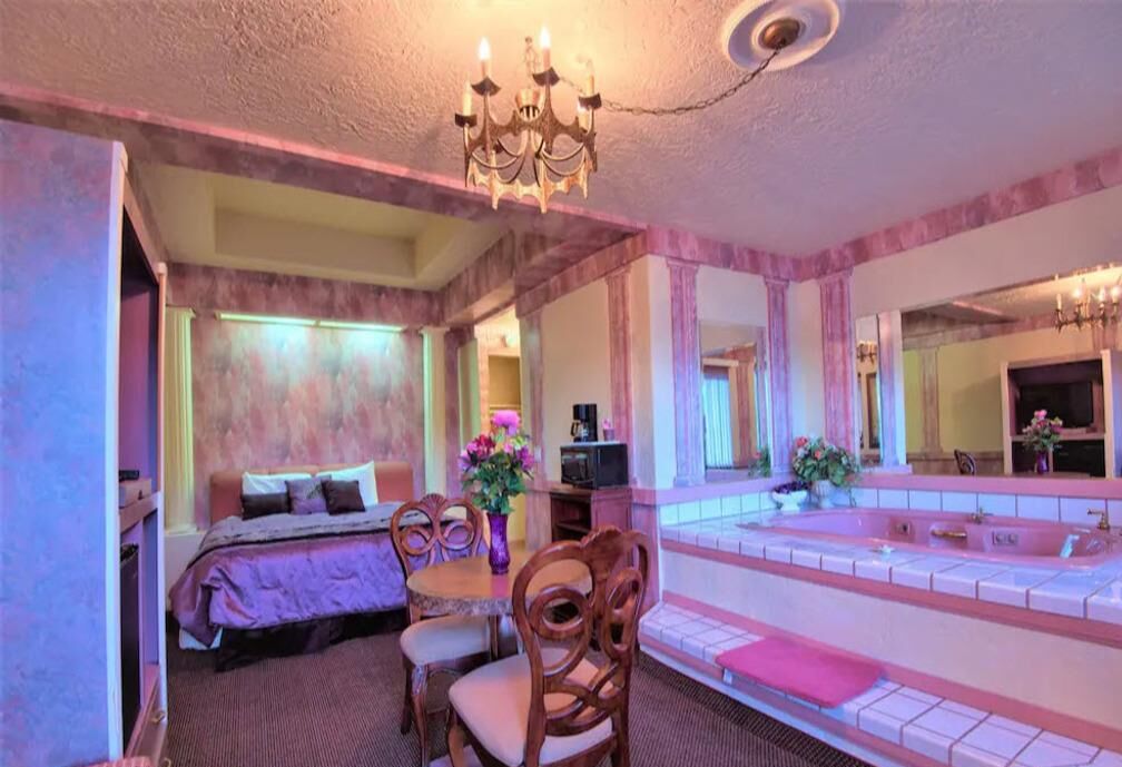 Une chambre d’hôtel comprenant un lit, un coin salon avec une table et des chaises, un grand miroir et une baignoire.