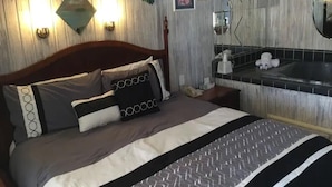 A hotel room with a wooden headboard, a bed with patterned bedding, a nightstand with a lamp, a wall-mounted mirror, and a bathroom with a sink and mirror.