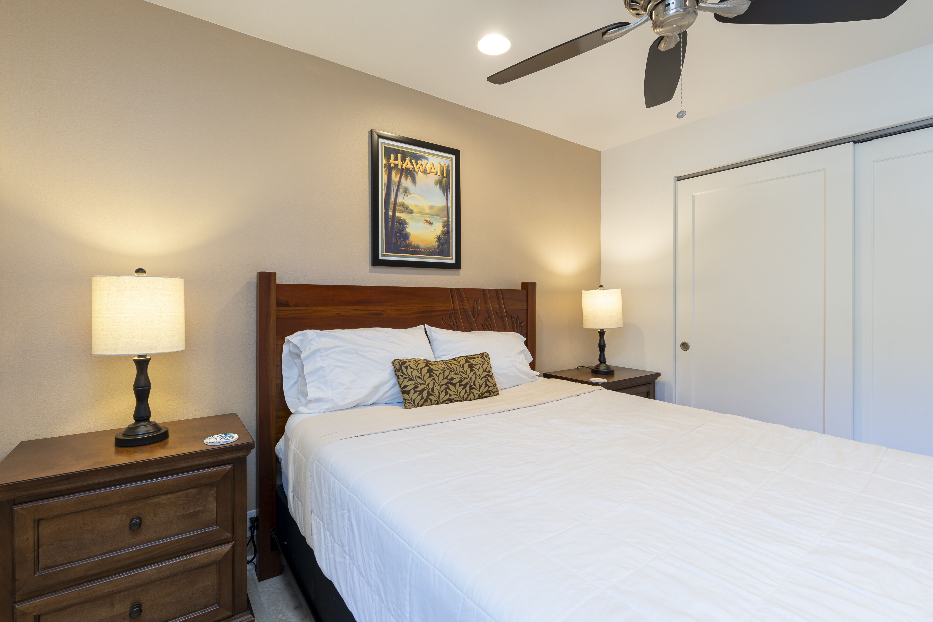 A neatly made bed with white linens, a wooden headboard, and a pillow with a leaf pattern. There is a framed picture of Hawaii on the wall above the bed.