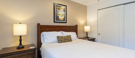 A neatly made bed with white linens, a wooden headboard, and a pillow with a leaf pattern. There is a framed picture of Hawaii on the wall above the bed.