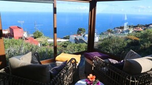 A balcony with wicker furniture, a small table with a vase of flowers, and a view of the sea and a coastal town.