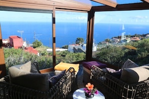 Varanda com mobiliário de vime, uma pequena mesa com um vaso de flores e vista para o mar e uma cidade costeira.