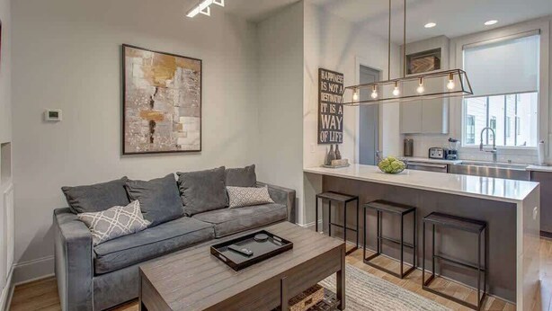 A modern living room with a grey sofa, a coffee table, and a kitchen area with a bar stool and pendant lighting.