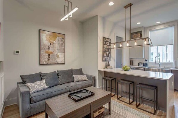 A modern living room with a grey sofa, a coffee table, and a kitchen area with a bar stool and pendant lighting.