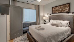 A hotel room with a bed, a ceiling fan, a TV mounted on the wall, a window with curtains, and a decorative wall piece.