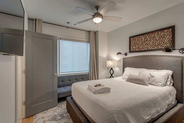 Une chambre d’hôtel avec un lit, un ventilateur de plafond, un téléviseur fixé au mur, une fenêtre avec des rideaux et un élément décoratif mural.