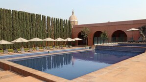 A swimming pool with lounge chairs and umbrellas, surrounded by a high wall of trees and a building with arches. - El Diezmo Hotel (San José Iturbide)