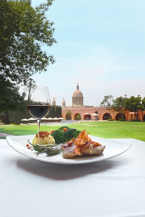 A dining table with a plate of food and a glass of red wine, set against a backdrop of a building with a dome and arches. - El Diezmo Hotel (San José Iturbide)