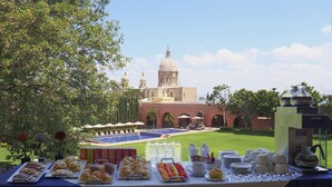 A buffet table with various food items and a coffee machine, set against a backdrop of a building with a dome and a pool. - El Diezmo Hotel (San José Iturbide)