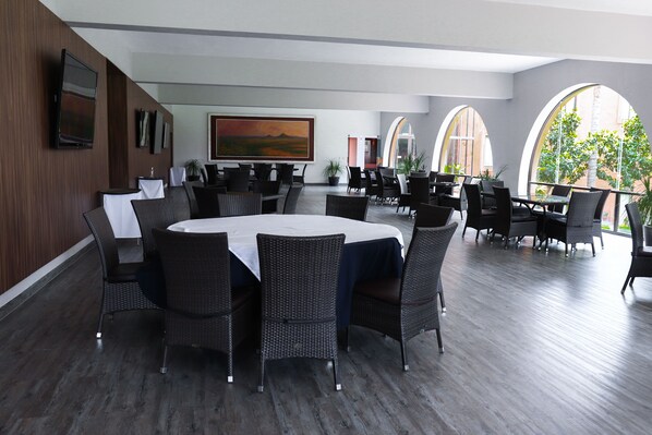A spacious indoor dining area with round tables, wicker chairs, and large windows offering views of the outdoors. - El Diezmo Hotel (San José Iturbide)