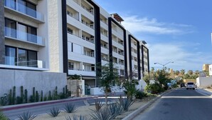 Un edificio moderno de varios pisos con balcones, una acera pavimentada y vegetación escasa.