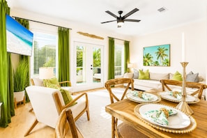 A room with a dining table set for four, a sofa, a ceiling fan, and a large window with green curtains.