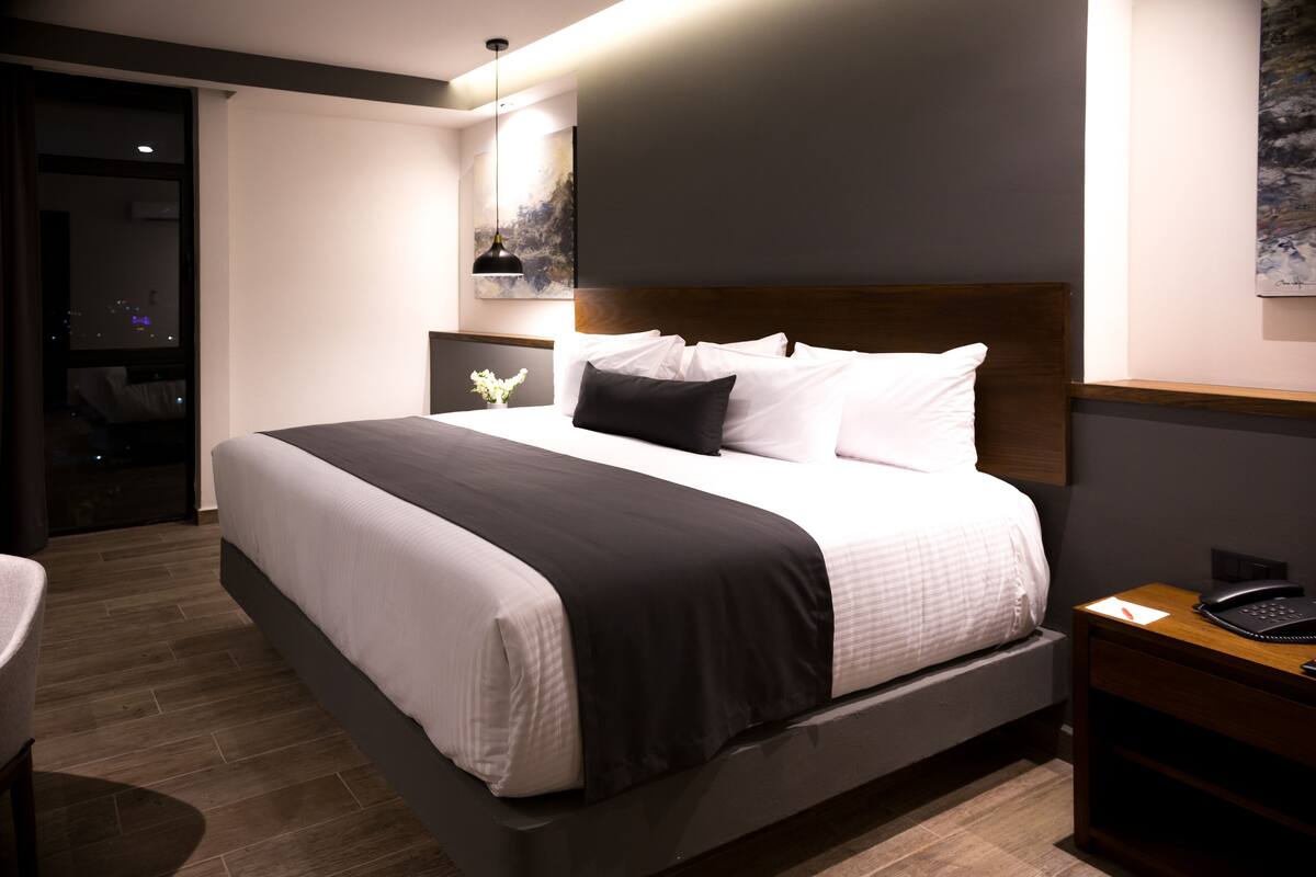 Een moderne hotelkamer met een groot bed, een nachtkastje met een lamp en uitzicht op de stad 's nachts.