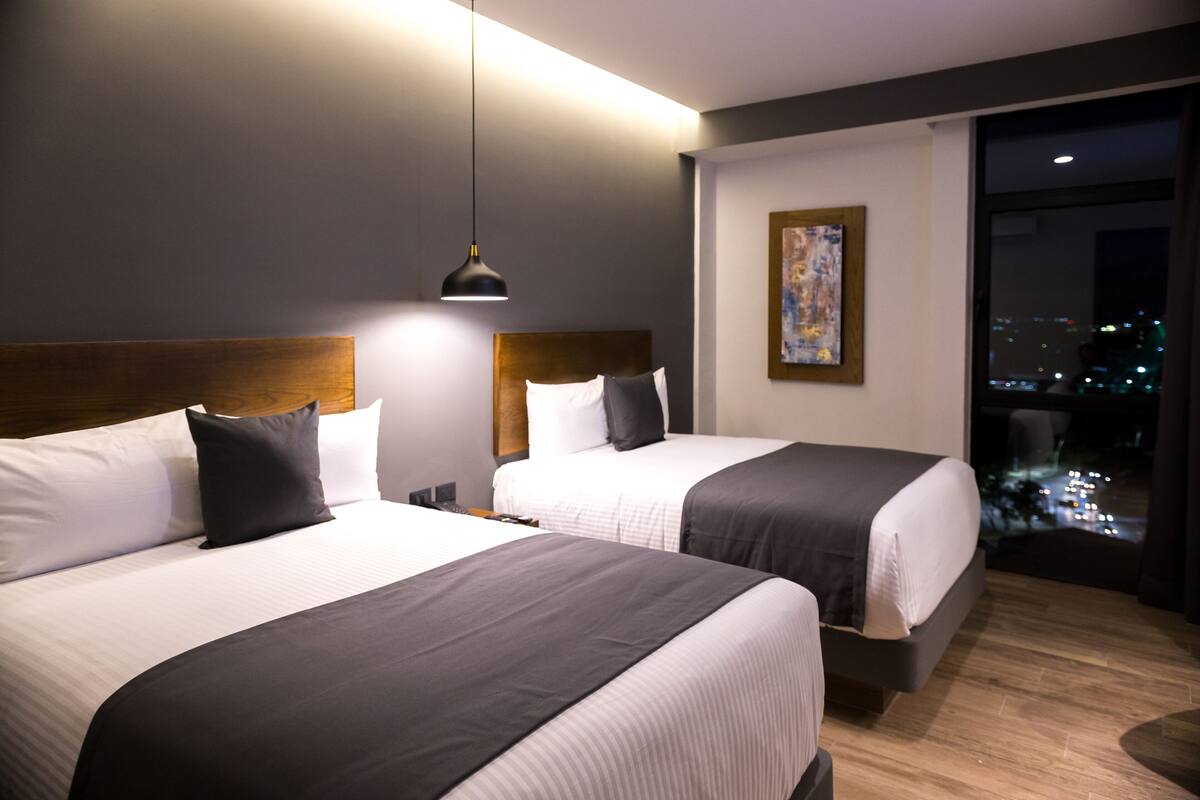 Een hotelkamer met twee bedden, een moderne hanglamp, een schilderij aan de muur en een uitzicht op de stad 's nachts.