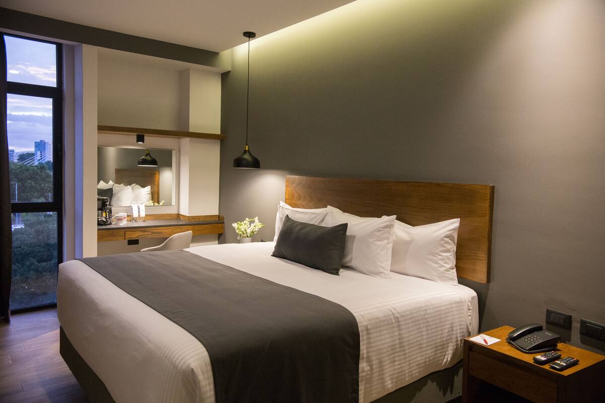 Een moderne hotelkamer met een groot bed, een nachtkastje met een telefoon en een uitzicht op de stad door een groot raam.