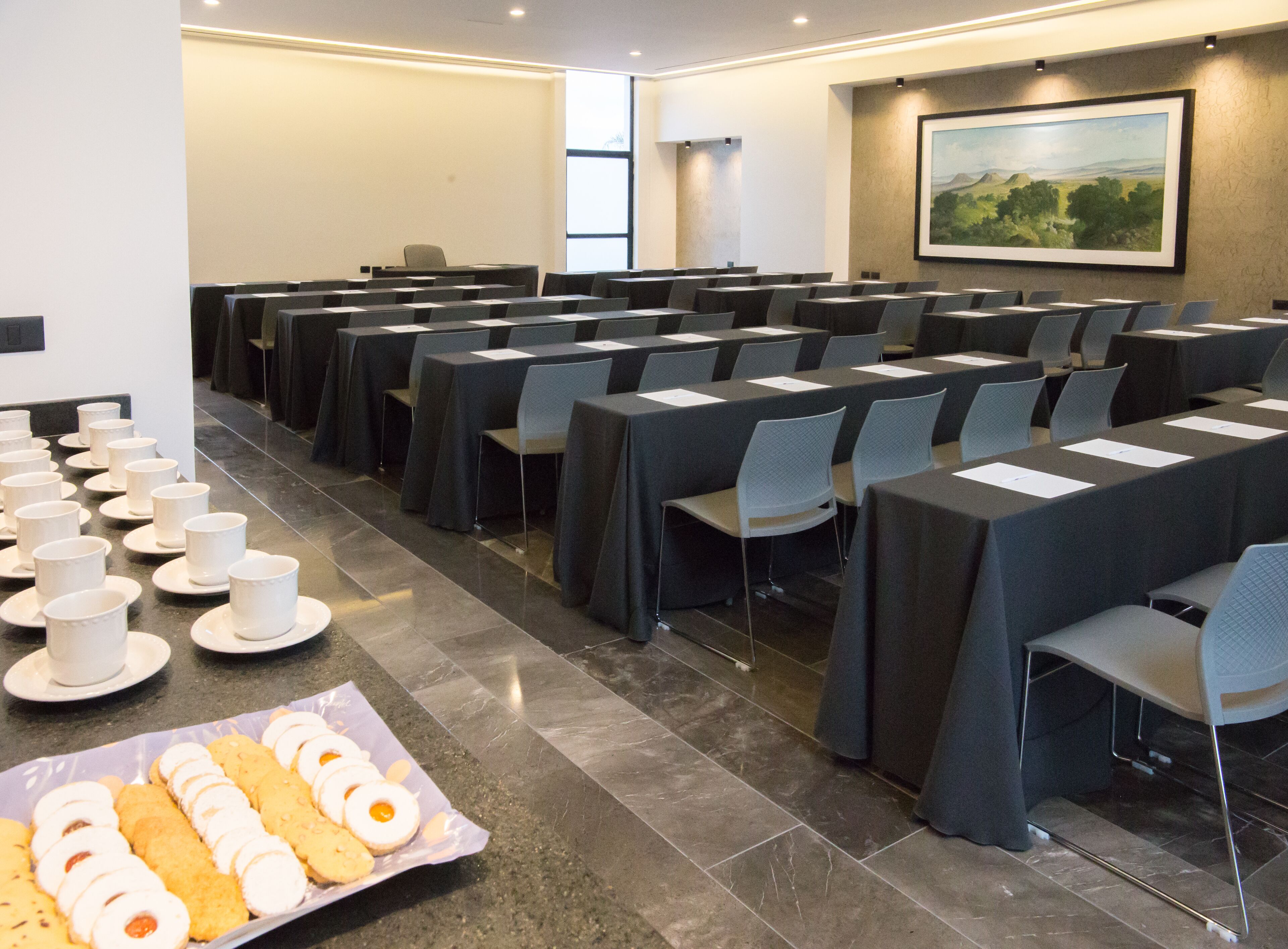 A conference room with rows of tables and chairs, a buffet table with pastries, and a painting on the wall.