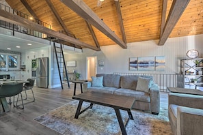 A spacious living area with a high ceiling, wooden beams, a sofa, a coffee table, and a kitchen in the background. - Airy Alpine Getaway w/ Lake Arrowhead Views! (Lake Arrowhead)