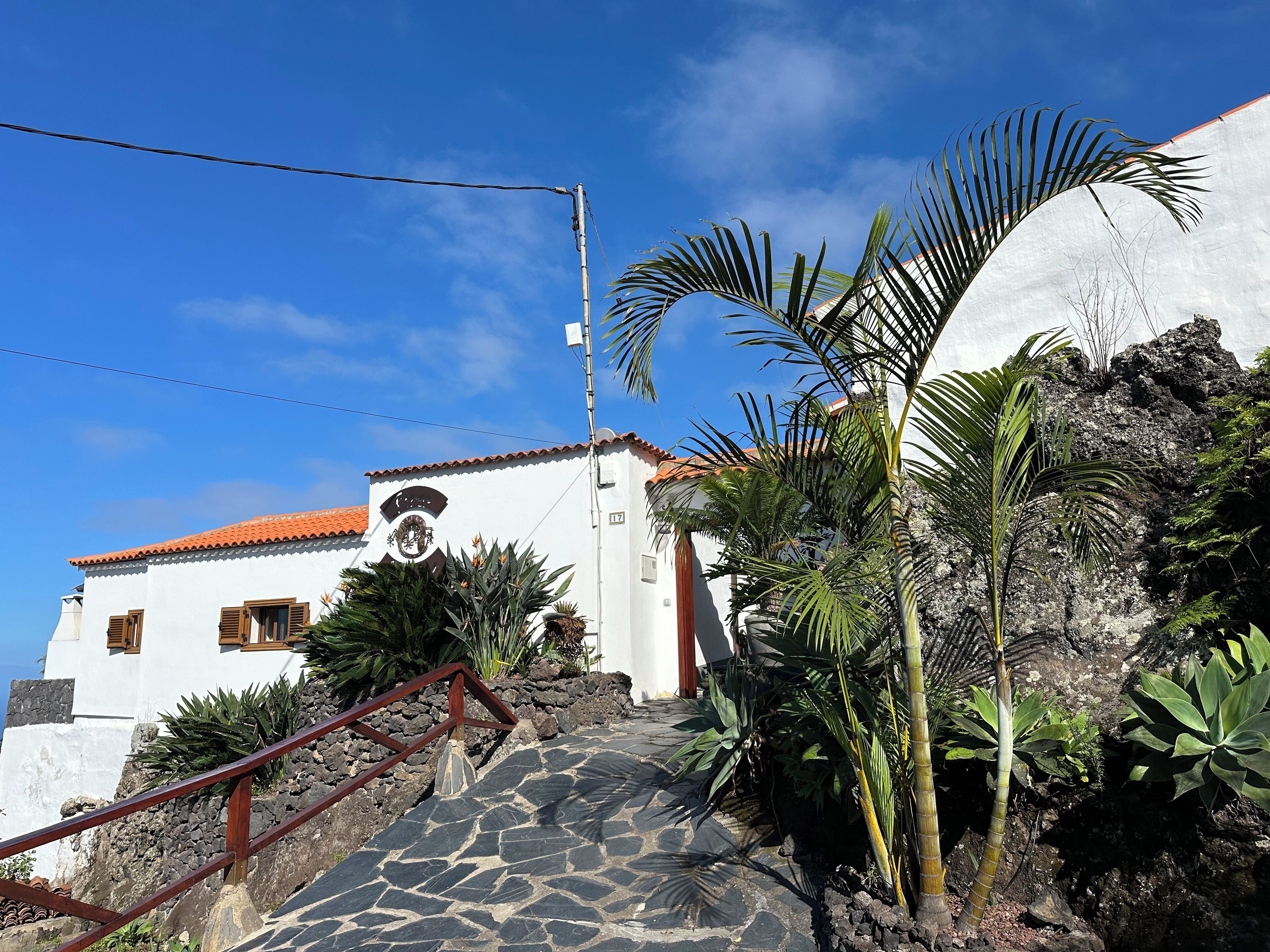 A white building with a red-tiled roof, a stone staircase, and tropical plants.