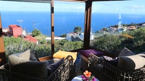 A balcony with wicker furniture, a small table with a vase of flowers, and a view of the sea and a coastal town.