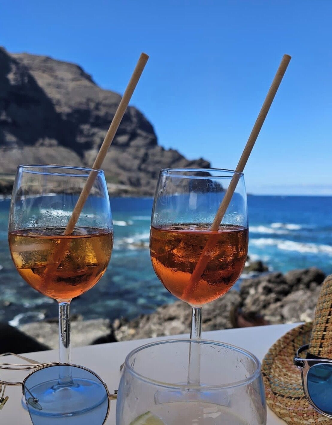 Dos vasos de una bebida color ámbar con pajitas, un par de gafas de sol y una cesta tejida sobre una mesa, con una costa rocosa y un cielo despejado al fondo.