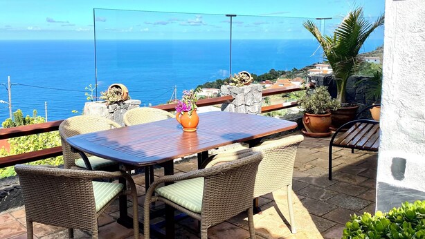 A patio with wicker furniture, a table, and potted plants, overlooking a coastal view.