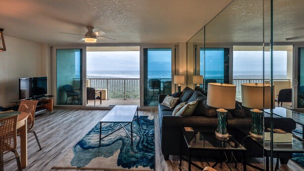 A modern living room with a sofa, coffee table, and TV, large windows with a view, and a ceiling fan. - Dazzling Dune Delight | Inlet #902 (New Smyrna Beach)