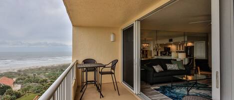A balcony with a table and chairs, a sliding glass door leading to a living area with a sofa and coffee table, and a view of the beach.